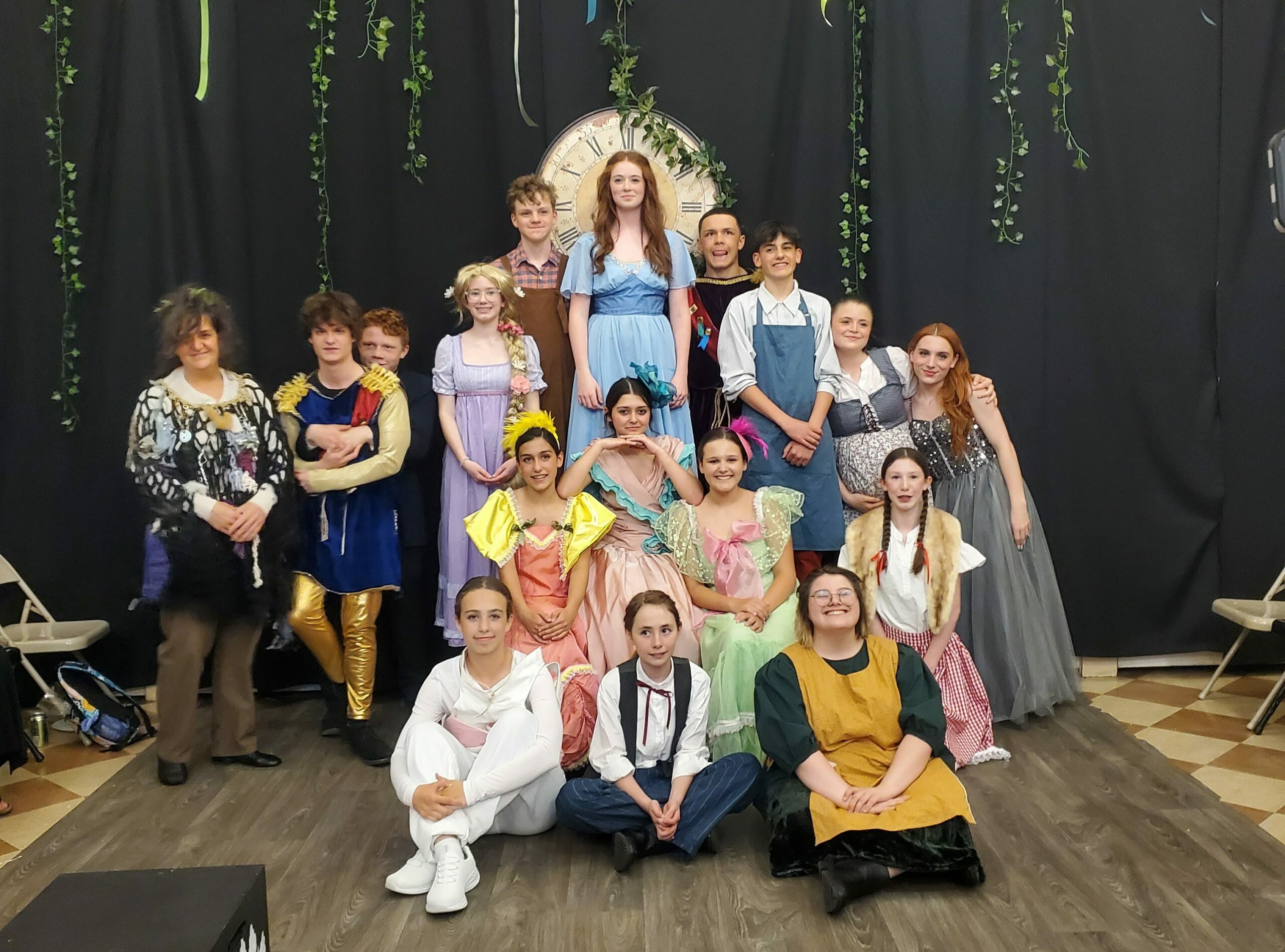 16 kids and one grown-up dressed in various fairy tale costumes such as Red Riding Hood, princes and princesses, and a cow pose in front of theater curtains and a huge clock